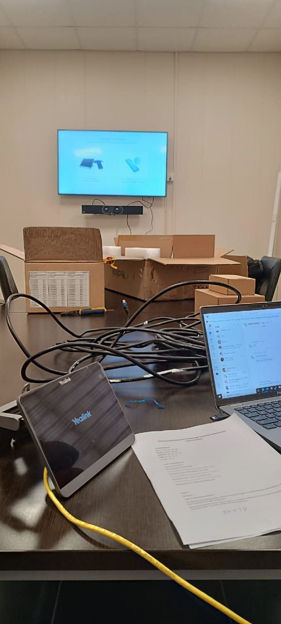 Une salle de réunion en cours d’installation audiovisuelle : écran mural et barre de son, kit de salle Yealink déballé, câblage et l’ordinateur portable d’un ingénieur affichant la fiche de montage.
