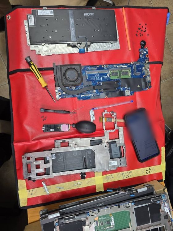 Un ordinateur portable professionnel réduit à ses composants sur un tapis antistatique de terrain lors d'une réparation sur site : carte mère avec sa mémoire, batterie, dissipateur et ventilateur, carte de stockage, ensemble écran et châssis.