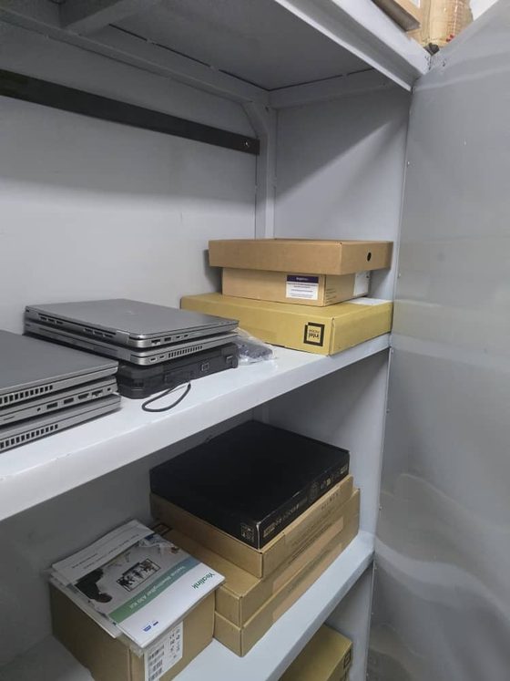 Staged IT hardware on warehouse racking: stacked notebooks, a small-form-factor desktop, a boxed conferencing room kit and spares held for scheduled jobs.